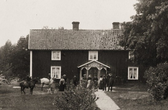 Svartvit bild av ett trähus med personer i gammaldags kläder utanför.