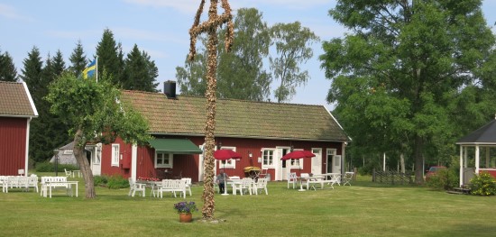 Rött trähus med vita cafémöbler och en vissen midsommarstång utanför.