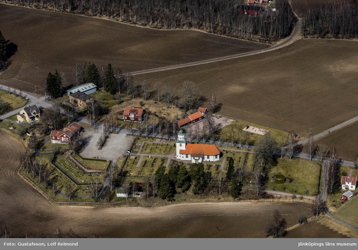 Flygfoto över kyrkan. Runt omkring ligger åkermark och lantliga hus.