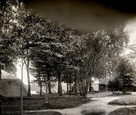 Gammal bild på en skog. I ena hörnet av bilden syns ett trätält.