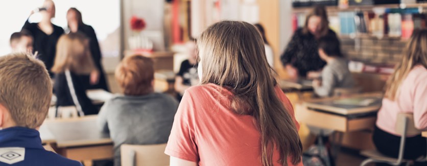 Ryggen på ett barn i rosa tröja som sitter i ett klassrum med andra barn